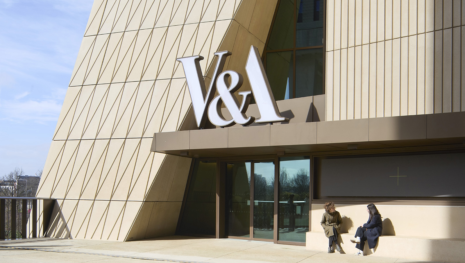 V&amp;A East Museum by O'Donnell+Tuomey in London, hero exterior of people sat outside the main entrance with the V&amp;A sign in view