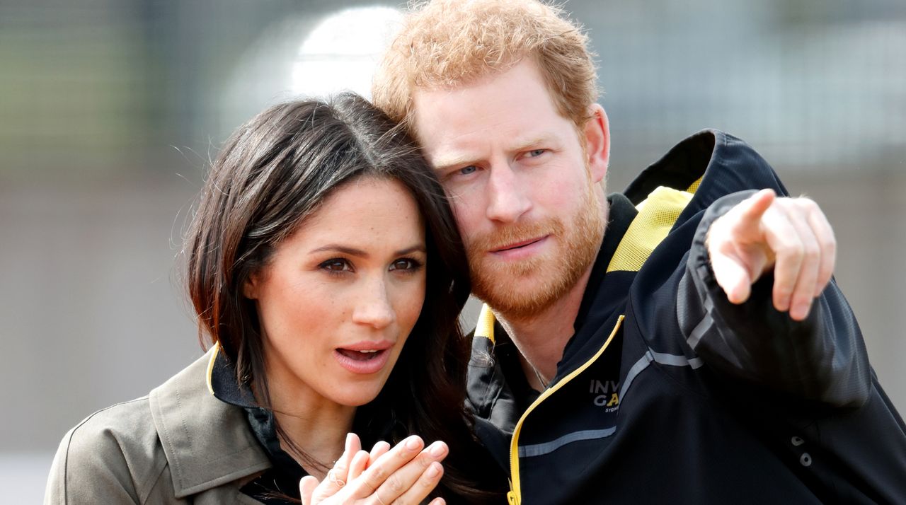 Prince Harry points out something to Meghan Markle, he is wearing a black raincoat with yellow detailing and Meghan is wearing a khaki green jacket