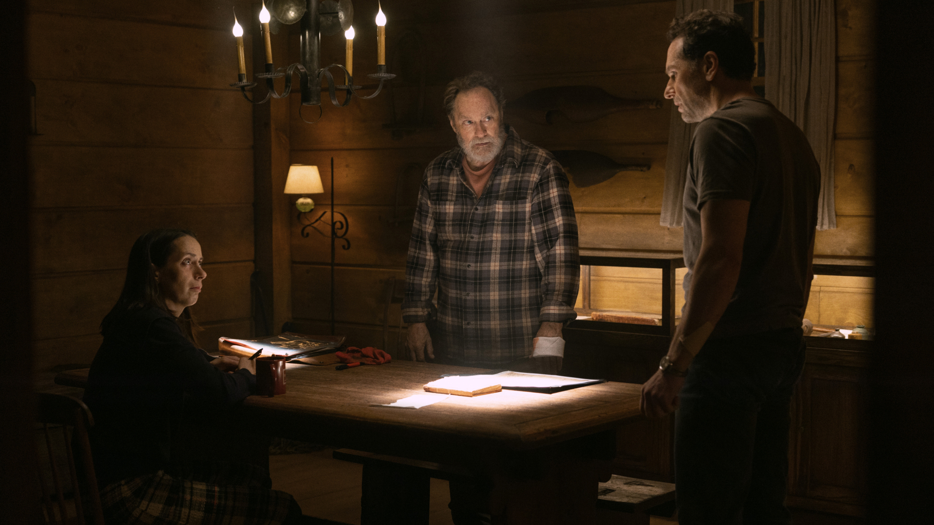 Three people gathered around a candlelit table. There are two men standing and one woman sitting.