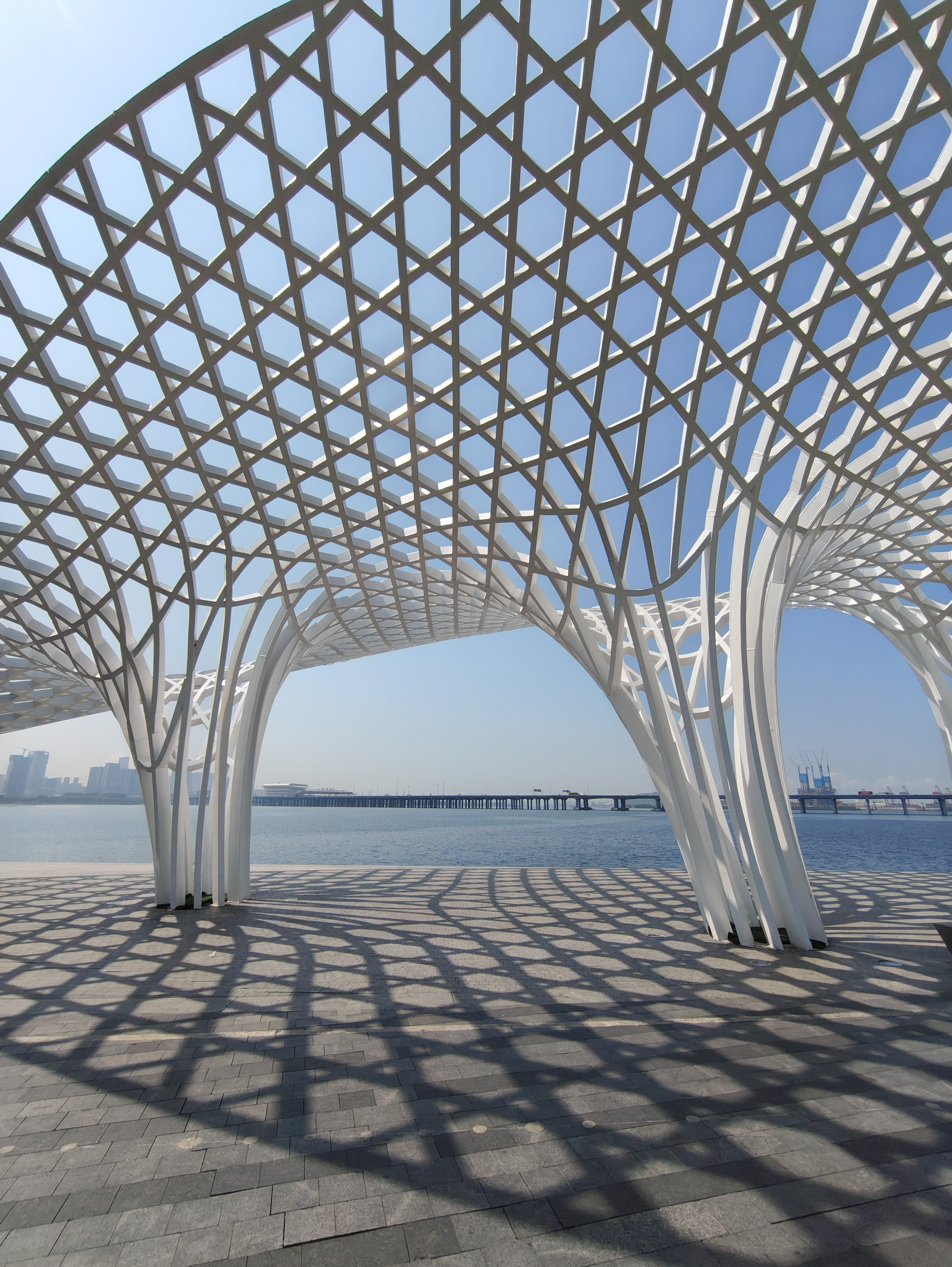 A wide-angle, low-angle shot of a large, white architectural canopy with a repeating diamond-shaped lattice pattern. The structure curves overhead like a giant wave, casting a dense, geometric shadow onto the paved ground. Through the openings of the structure, a calm bay, a long bridge, and a distant city skyline are visible under a bright, clear sky.