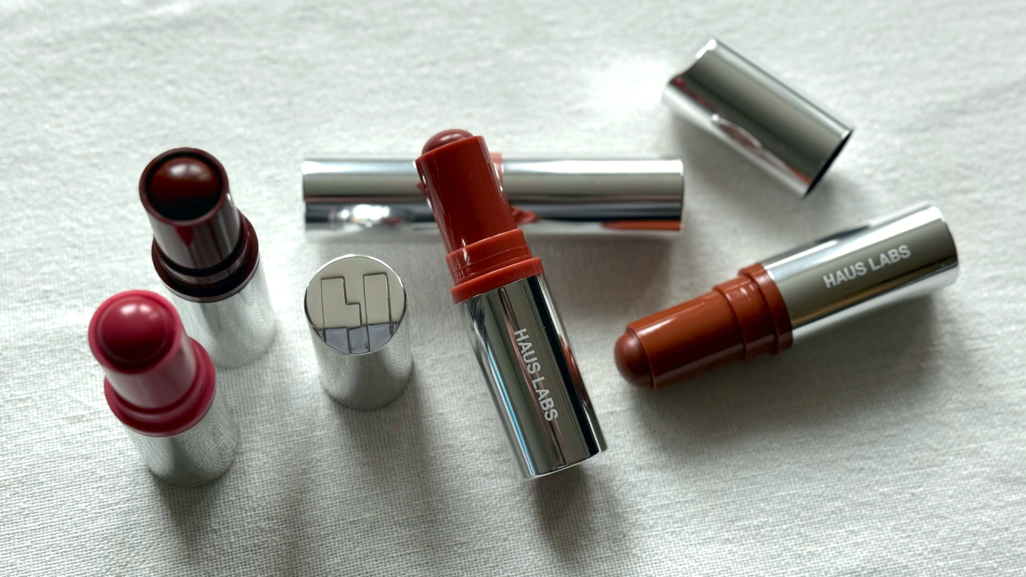 Bird's eye view of five Haus Labs Glassy Blush Balm Sticks, four with their lids off, arranged in a still life ensemble on an off-white linen background