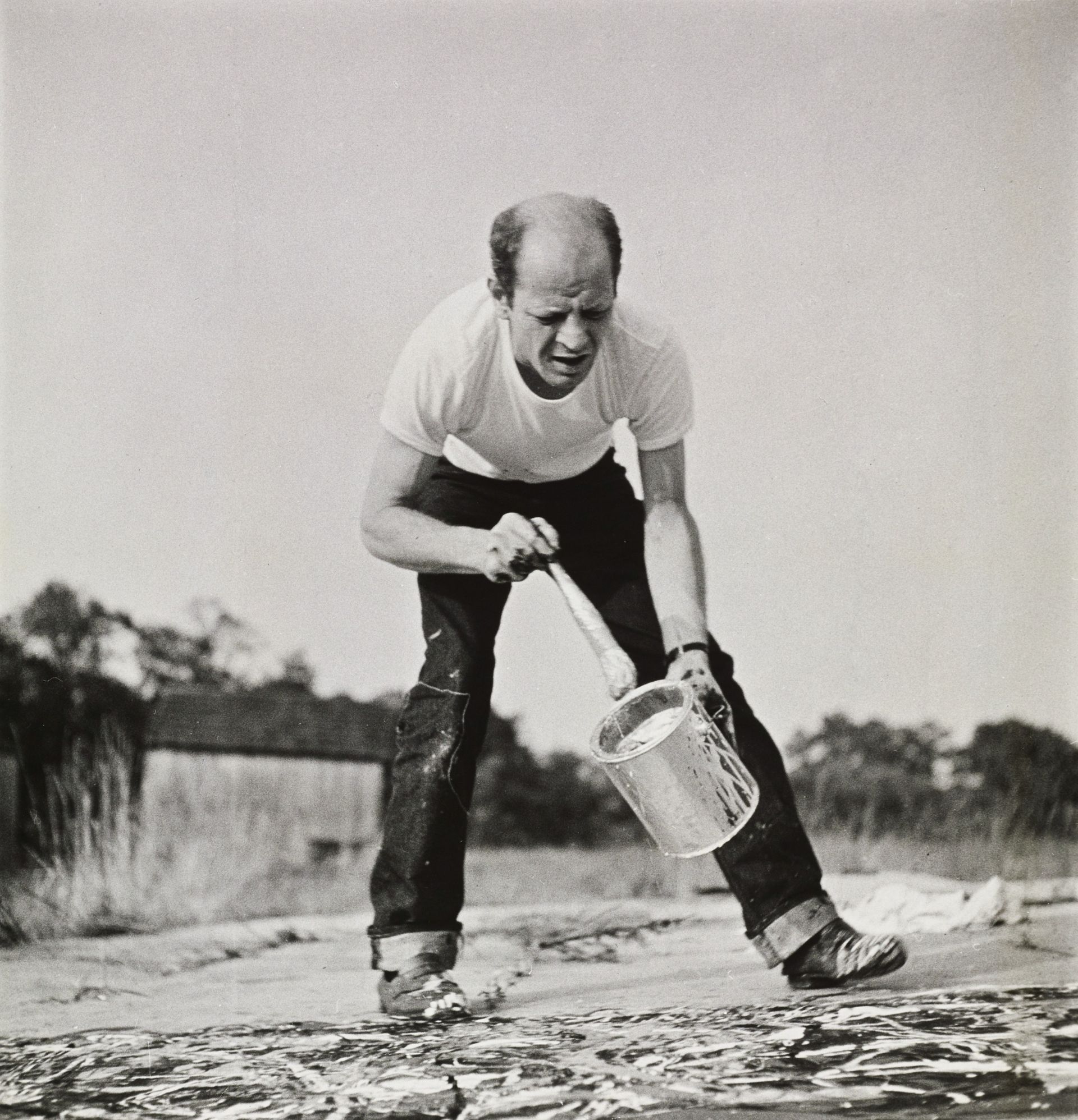 Black-and-white photograph of Jackson Pollock bending over to paint a canvas that's lying on the floors