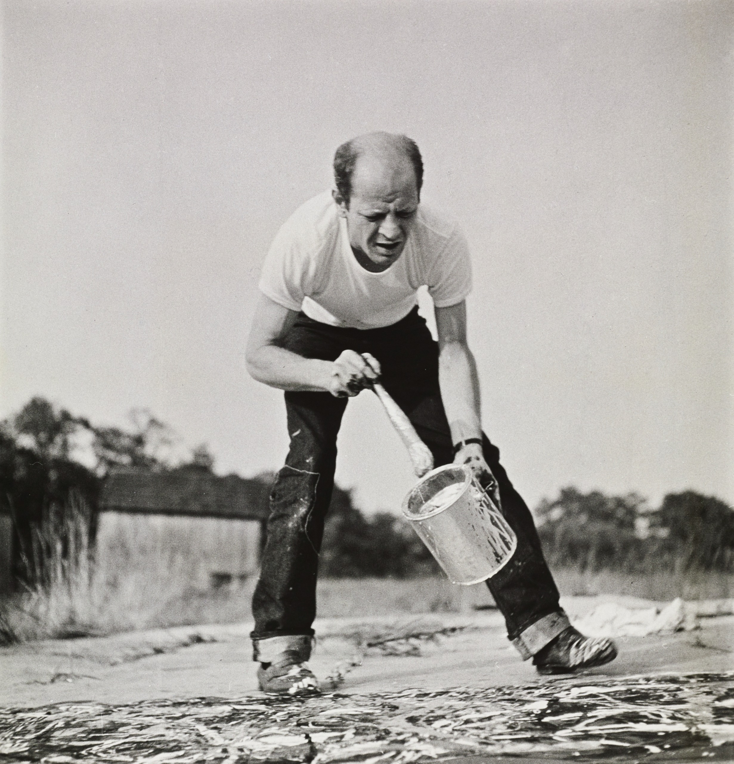 Black-and-white photograph of Jackson Pollock bending over to paint a canvas that's lying on the floors