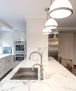 A marble kitchen island with a silver sink and two faucets, three curved pendant lights above it, and a pillar,cabinets, and silver ovens beyond it