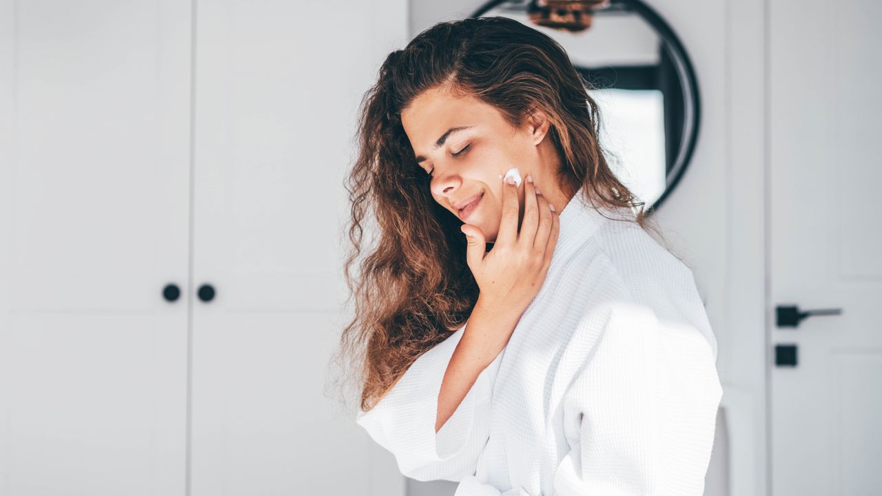 Young woman with long curly hair in bathrobe enjoys daily routine applying moisturizing cream on face in bedroom at home closeup mirror reflection