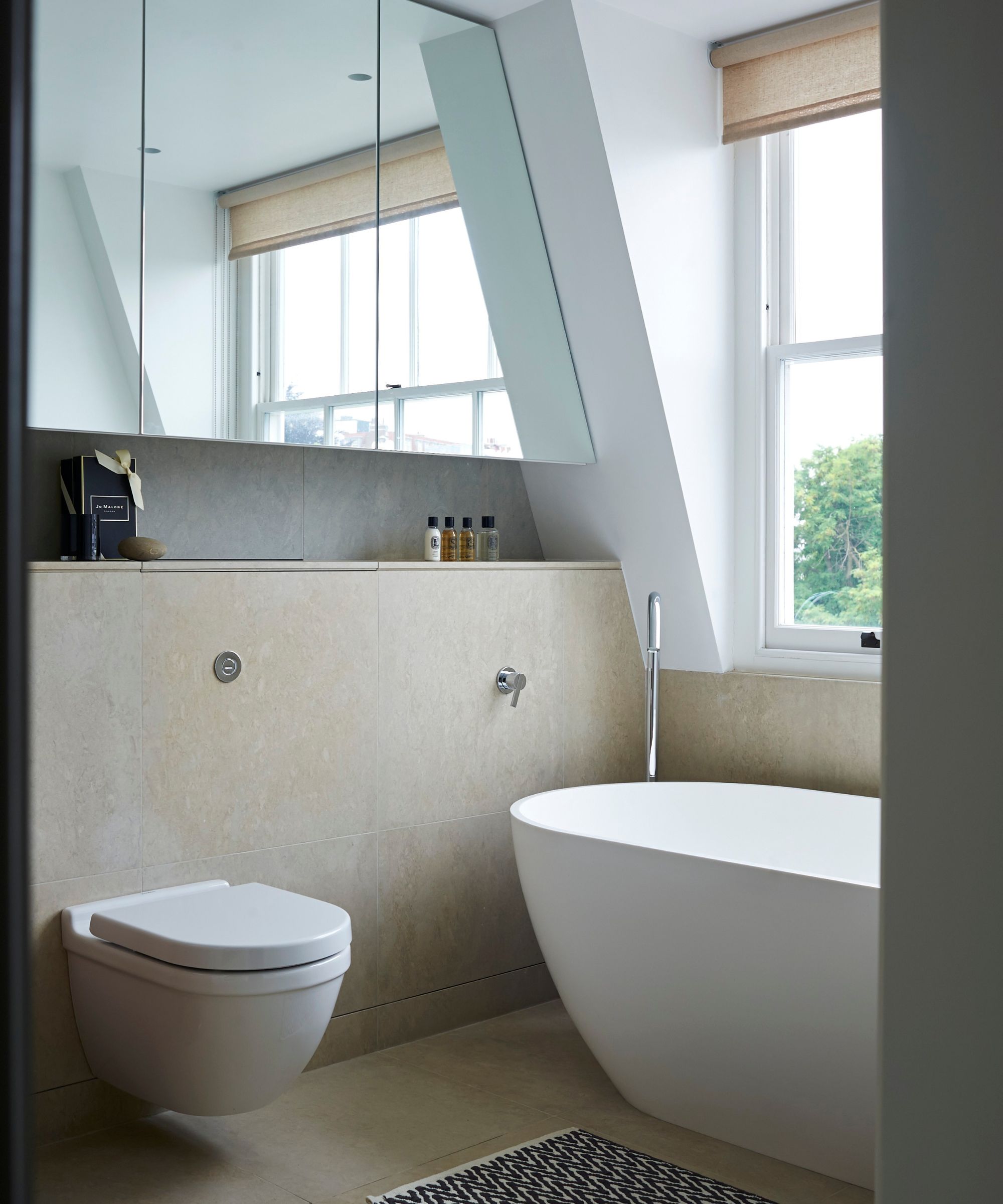 A bright bathroom with neutral tiles, large white bathtub, blind, mirror and Jo Malone toiletries.