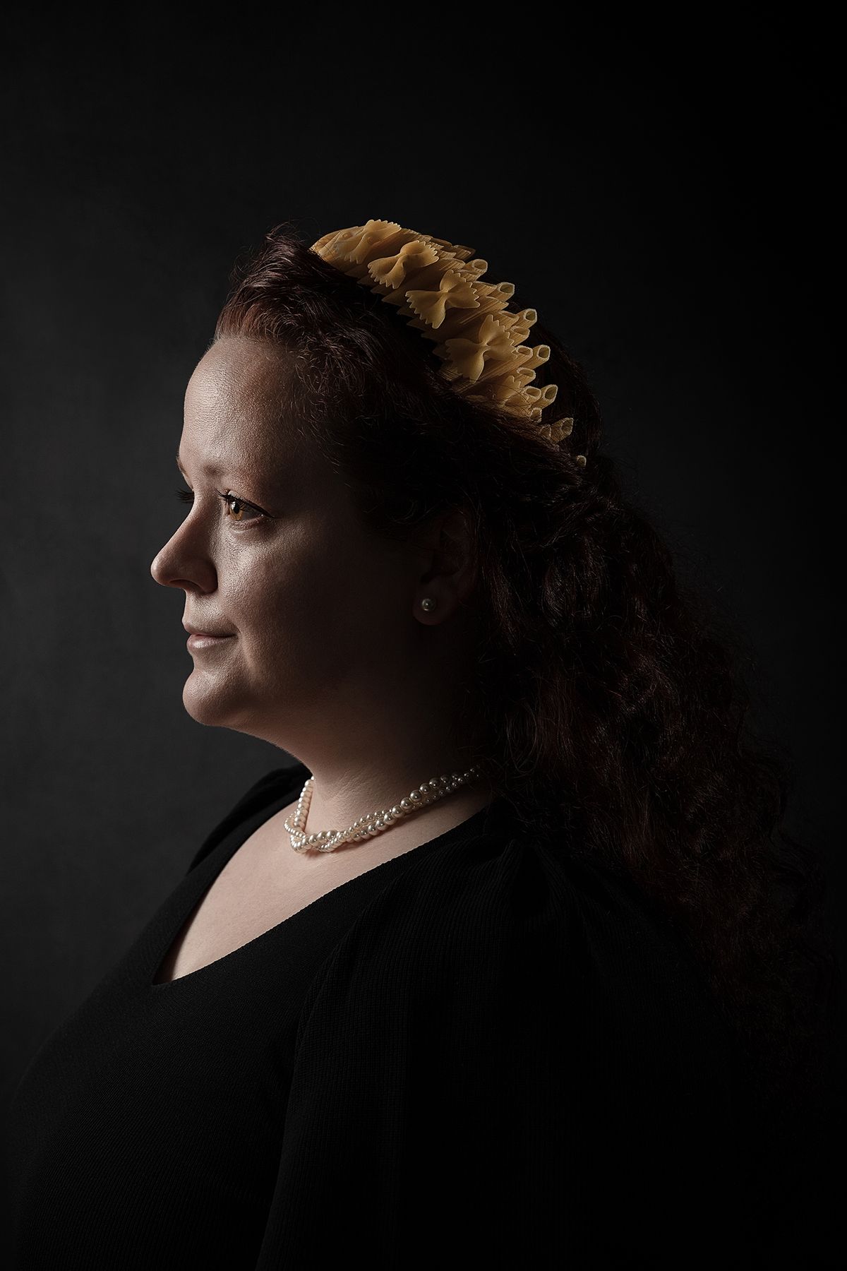 A woman with curly hair wears a headband made from raw pasta, against a dark, dramatic background