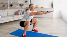 man in a white tshirt, black shorts and red trainers performing a squat on a blue mat on wooden floors in a living room. there's a sofa and shelving unit behind him. 