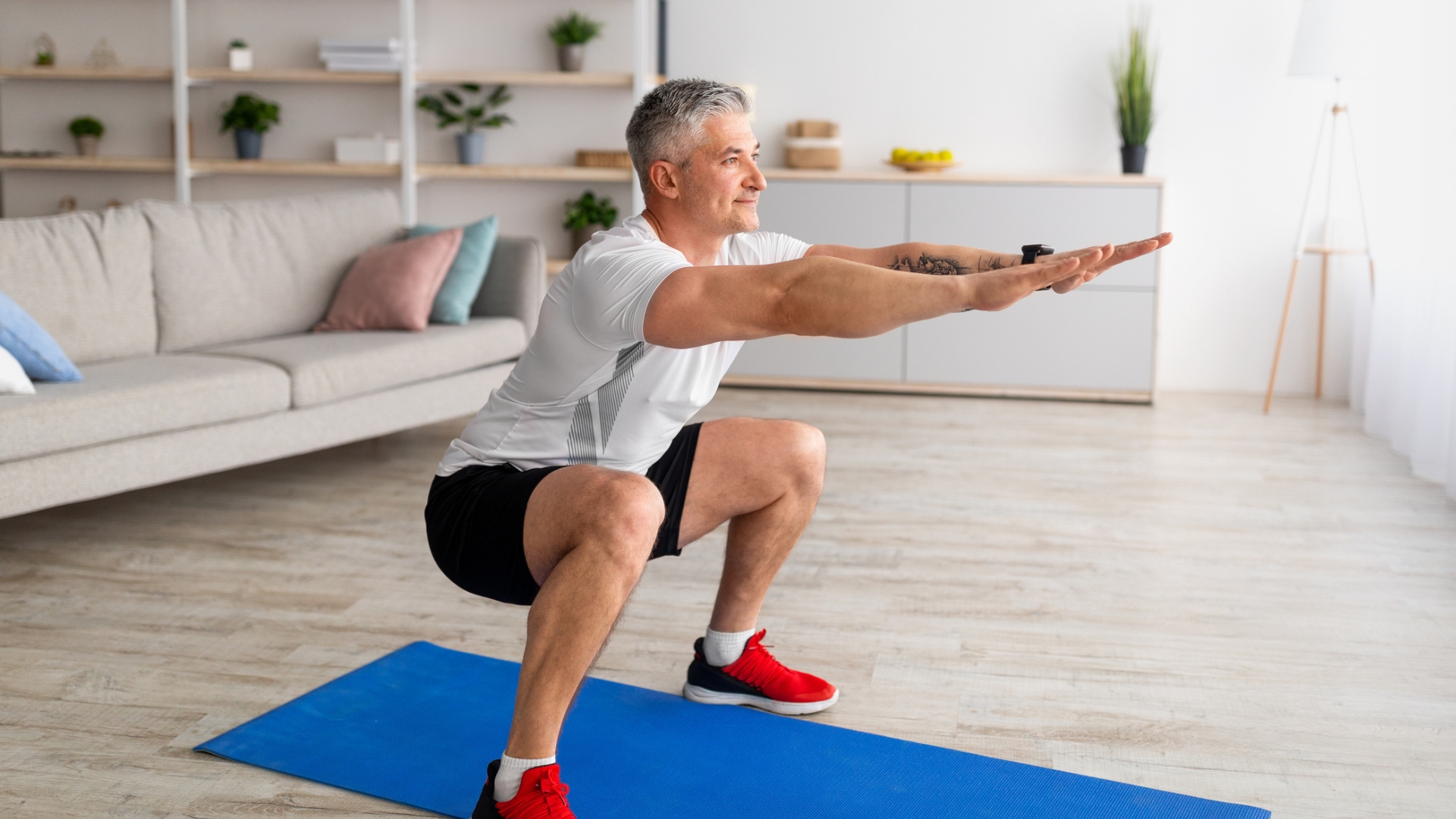 man in a white tshirt, black shorts and red trainers performing a squat on a blue mat on wooden floors in a living room. there's a sofa and shelving unit behind him.