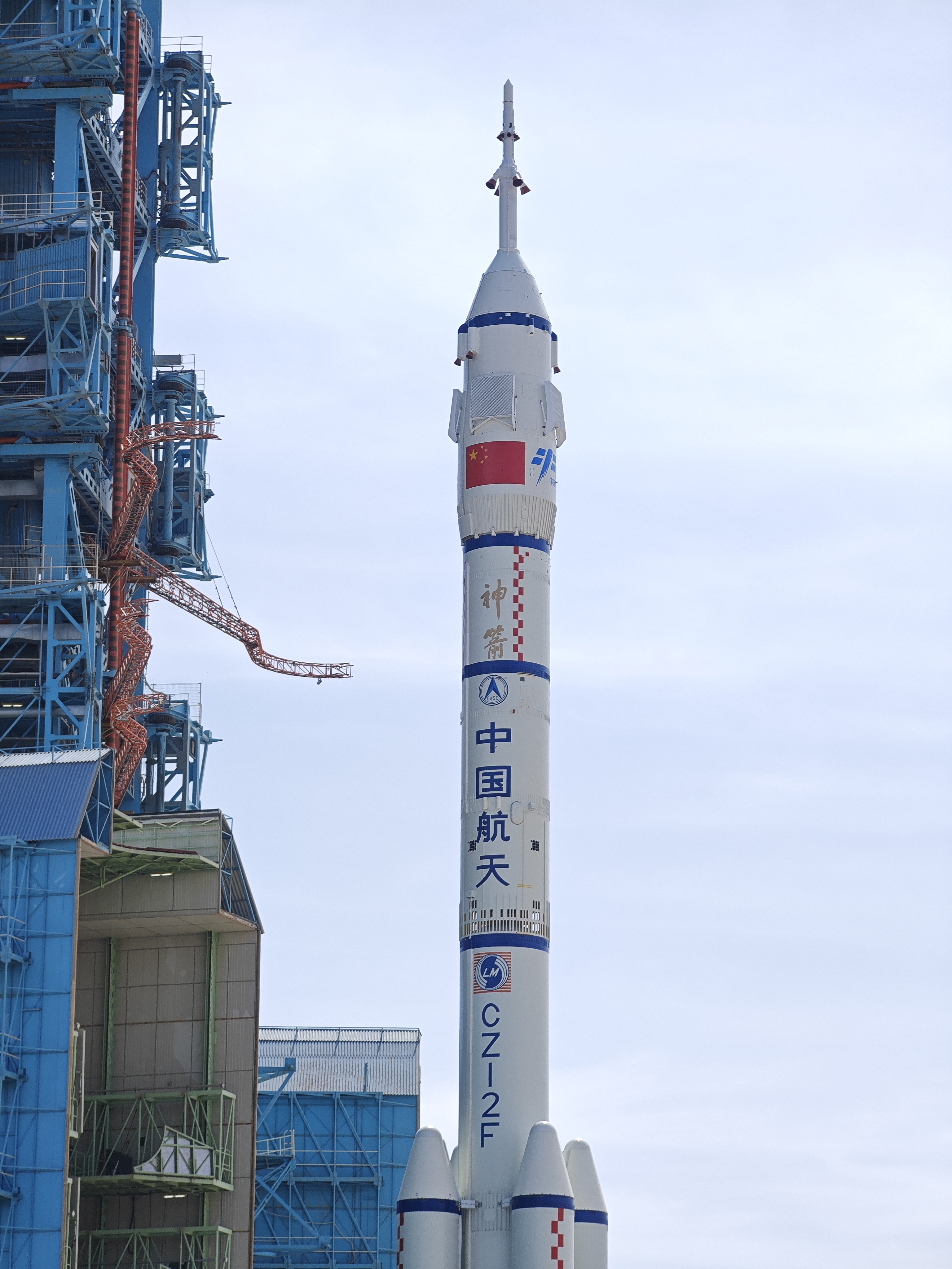 Duża biała rakieta z czterema małymi dopalaczami wtacza się na platformę startową otoczona czerwonymi flagami z żółtymi chińskimi znakami