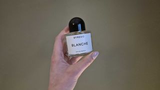 A close up Digital Beauty Writer, Naomi Jamieson holding a glass bottle of Byredo's Blanche eau de parfum, in front of a green background