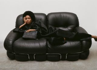 A black model with Afro laying on a black leather sofa with a black crocodile-pattern handbag against a grey background