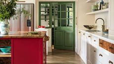 White kitchen with white cabinets, red kitchen island with wood countertop, wood flooring, and green door leading to pantry