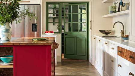 White kitchen with white cabinets, red kitchen island with wood countertop, wood flooring, and green door leading to pantry