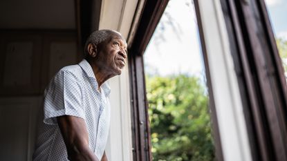 An older man looks out the window at home.