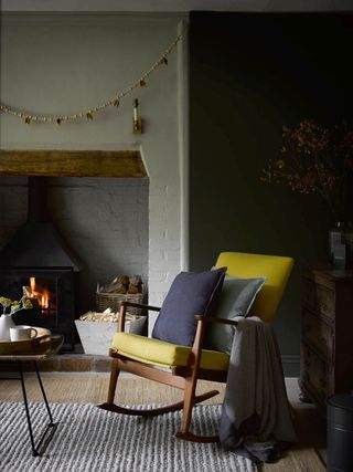 winter decor ideas, cozy living room with rocking chair in front of log burner, chunky rug, coffee table, logs, cushions and blankets, winter decoration above fireplace, wall light