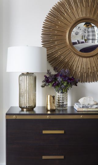 Chest of drawers in dark wood with gold trim, table lamp, vases and books, and gold framed mirror above