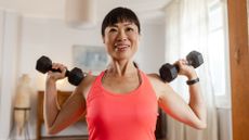 Woman holding two dumbbells against her shoulders, standing in living room after asking are free weights better than machines