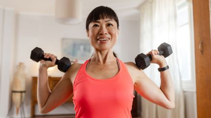 Woman holding two dumbbells against her shoulders, standing in living room after asking are free weights better than machines