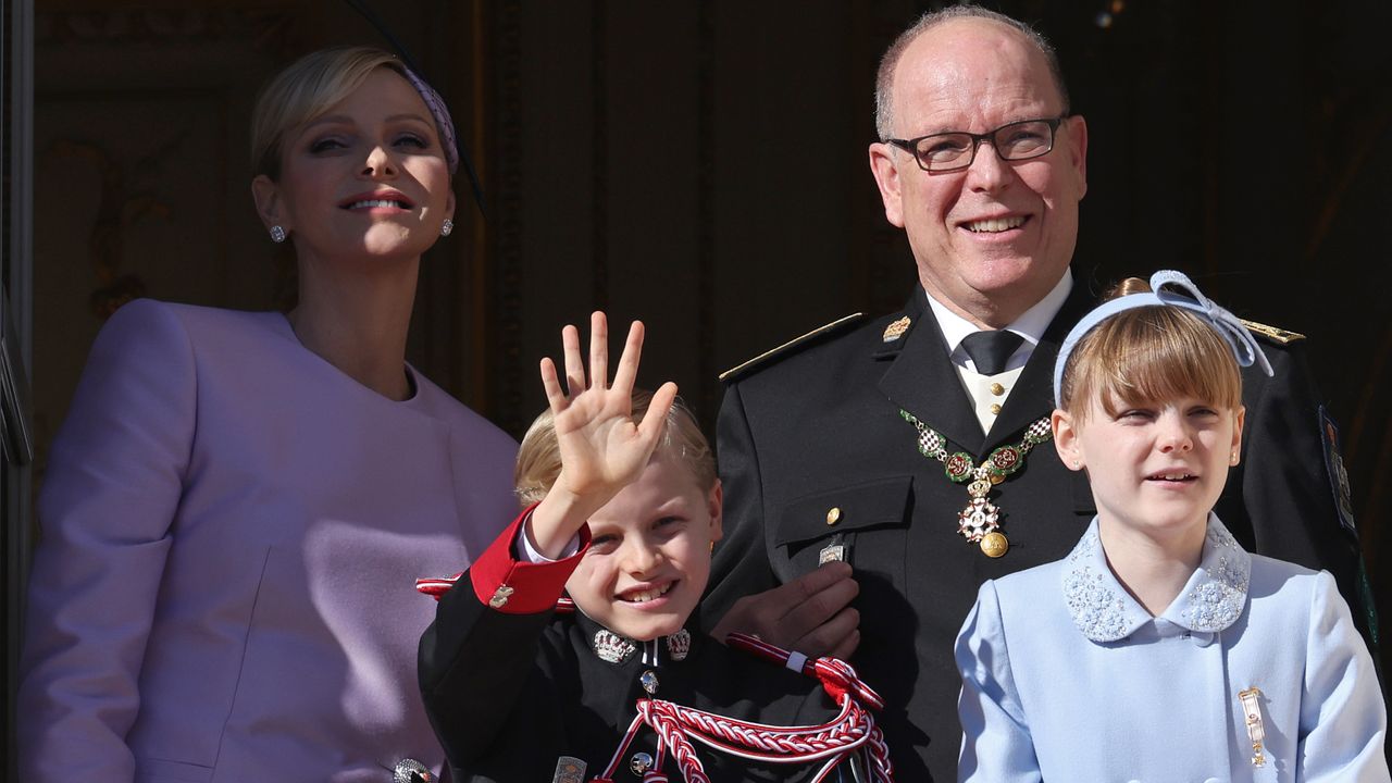 Princess Charlene, Prince Albert, Princes Jacques and Princess Gabriella wearing purple coats and military uniforms waving on a sunny balcony