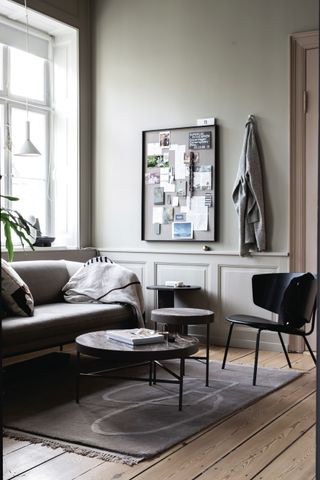 A living room by Nest using a grey wall paint shade with green undertones, grey sofa, noticeboard, coat on hook, black chair, set of three coffee tables and grey rug with abstract detail