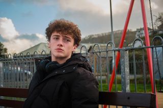 Jaiden (Austin Taylor) sits on a bench next to a park, wearing a winter coat, with headphones around his neck. He has a sad look on his face.