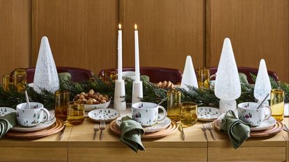 Christmas table setting on bare wooden table with glass trees, greenery, candles and coloured yellow glasses 