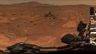 view of a rocky patch of reddish mars ground, with a mountain in the background and a portion of nasa's perseverance mars rover in the foreground.
