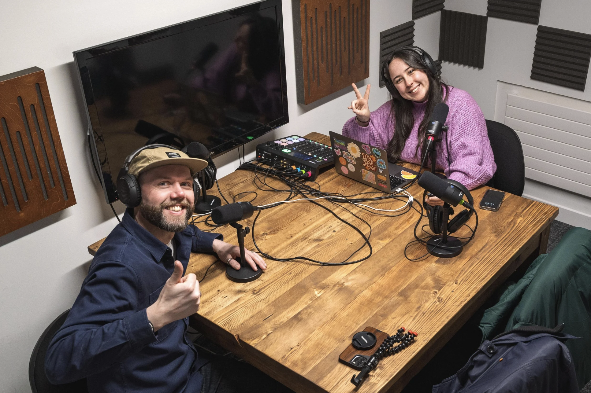 A man giving a thumbs-up and a woman flashing a peace sign smile from a wooden table equipped with microphones, headphones, and a mixing console in a podcast studio.