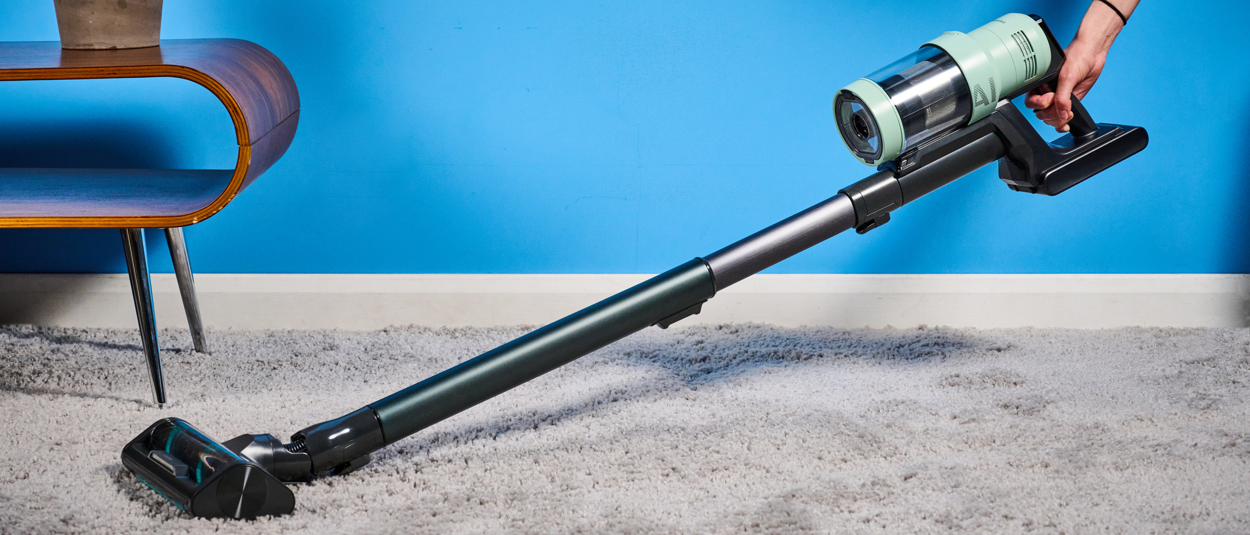 the samsung bespoke jet AI vacuum cleaner in mint green against a blue background, showing its ability to clean carpets and baseboards, as both a stick and a handheld
