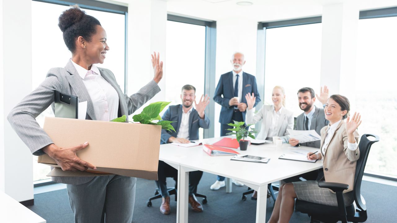 A female executive walks out of the office with a box of her belongings while co-workers wave goodbye behind her.