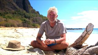 Phillip Schofield sits alone, wearing a grey t-shirt and looking dishevelled, on an uninhabited tropical island beach in Phillip Schofield Cast Away 2024 on Channel 5