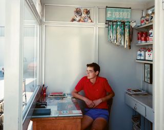 A caucasian man wearing a red polo shirt and blue shorts sitting in a room with his right elbow resting on a wooden table looking outside the white framed window. The table has stationeries and books laid on it. behind him are 3 floating shelves and a floating drawer with a silver knob. The 3 floating shelves are occupied with white and red bottles.