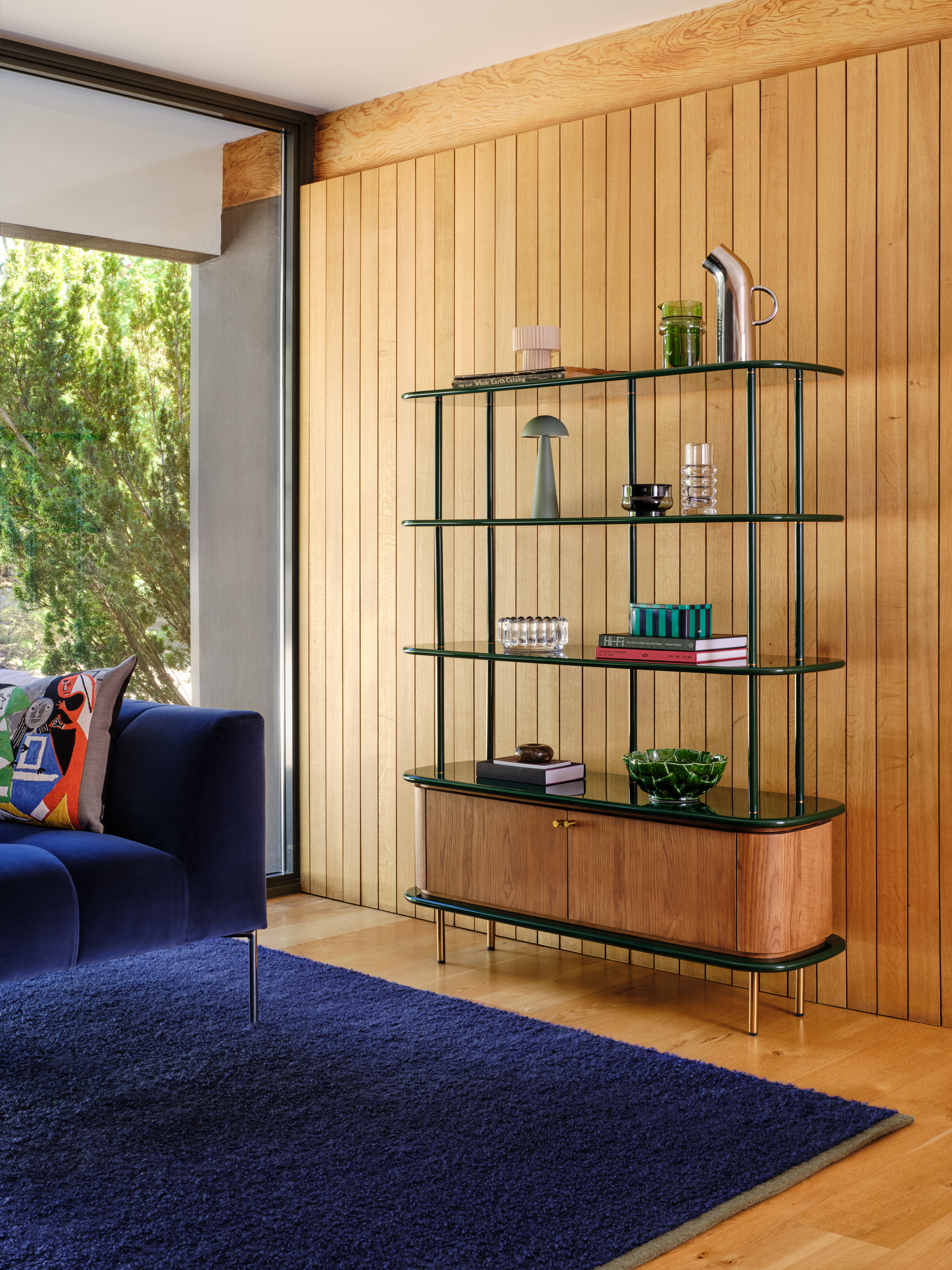 Image of a wooden console unit with dark green lacquer shelving. The piece is sitting in a living room with wood-paneled walls and a dark navy sofa and rug.