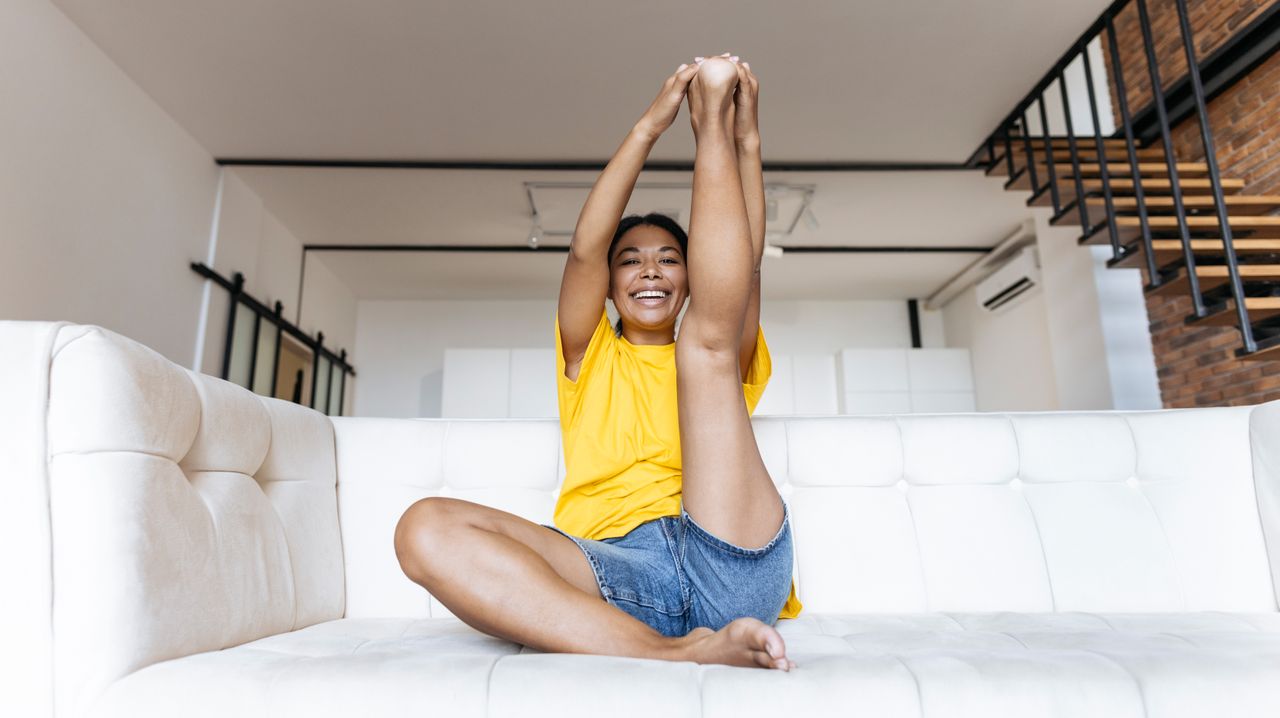 A woman sits on a couch stretching her leg in the air. Her right leg is folded underneath her and her left leg is pointing towards the ceiling; her hands are clasped around the sole of her foot. Behind her we see a wooden staircase.