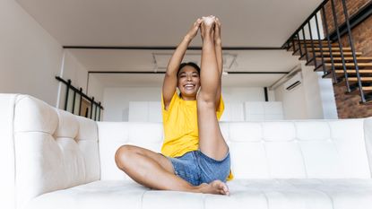 A woman sits on a couch stretching her leg in the air. Her right leg is folded underneath her and her left leg is pointing towards the ceiling; her hands are clasped around the sole of her foot. Behind her we see a wooden staircase.