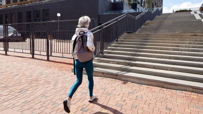 Woman wearing exercise clothes and backpack, walking towards concrete staircase with rail outdoors, representing how far should you walk every day