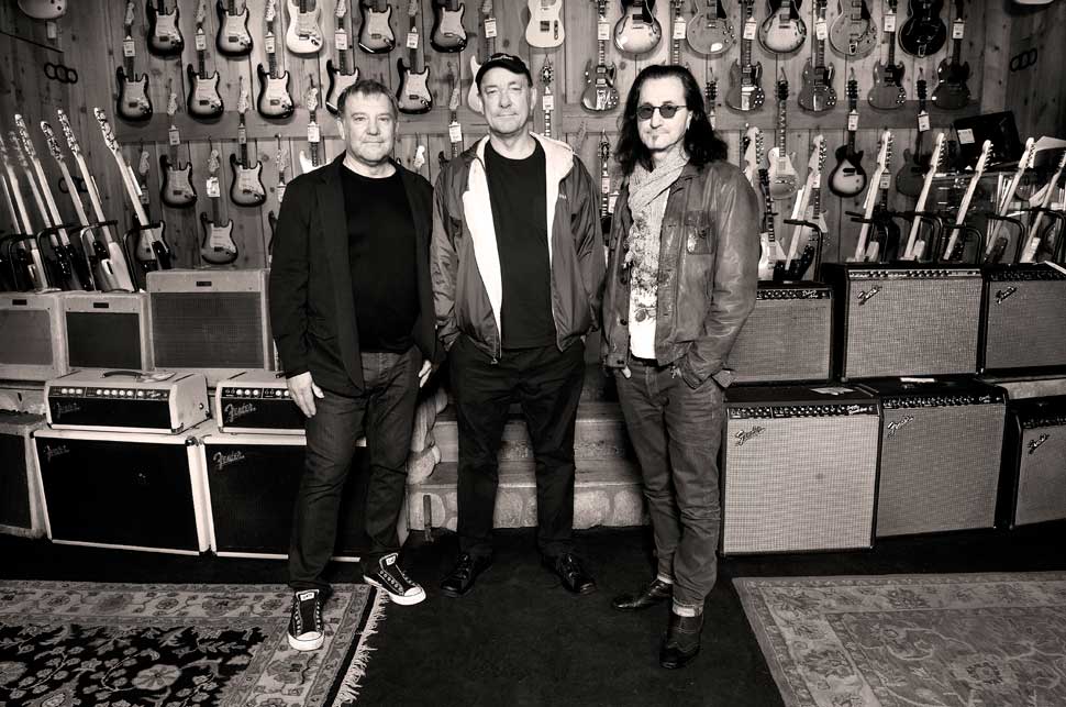 Rush standing in front of rows of guitars and amplifiers