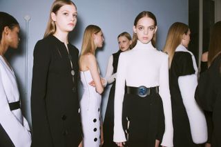 Model wears a white turtle-neck top with a black skirt and plexiglass belt, others wear monochrome overcoats