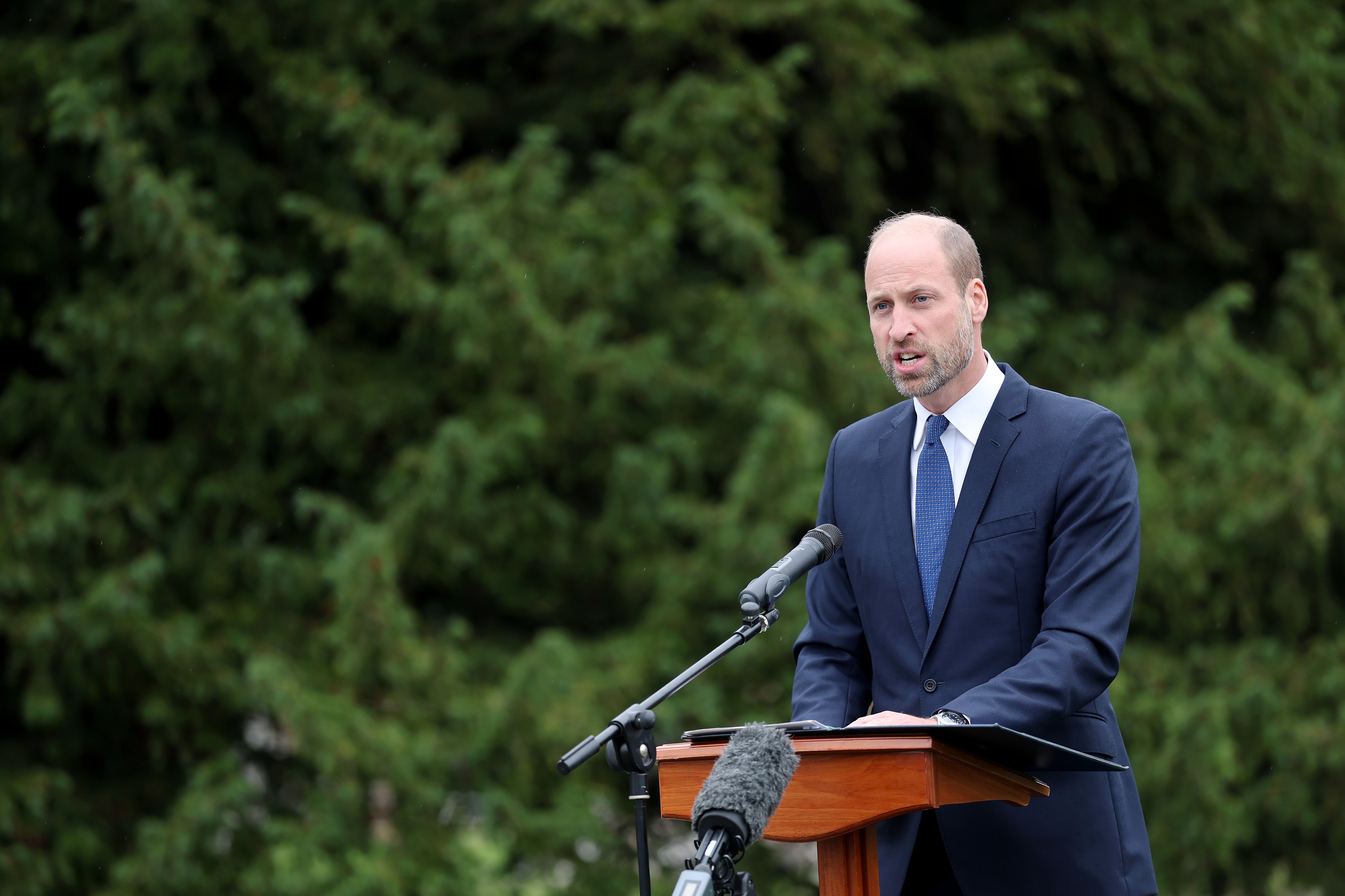 Prince William wearing a blue suit giving a speech in front of trees