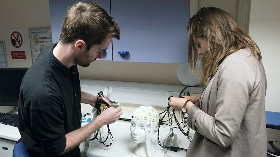 two light-skinned people, a man and a woman, place an electrode-covered cap on a mannequin head