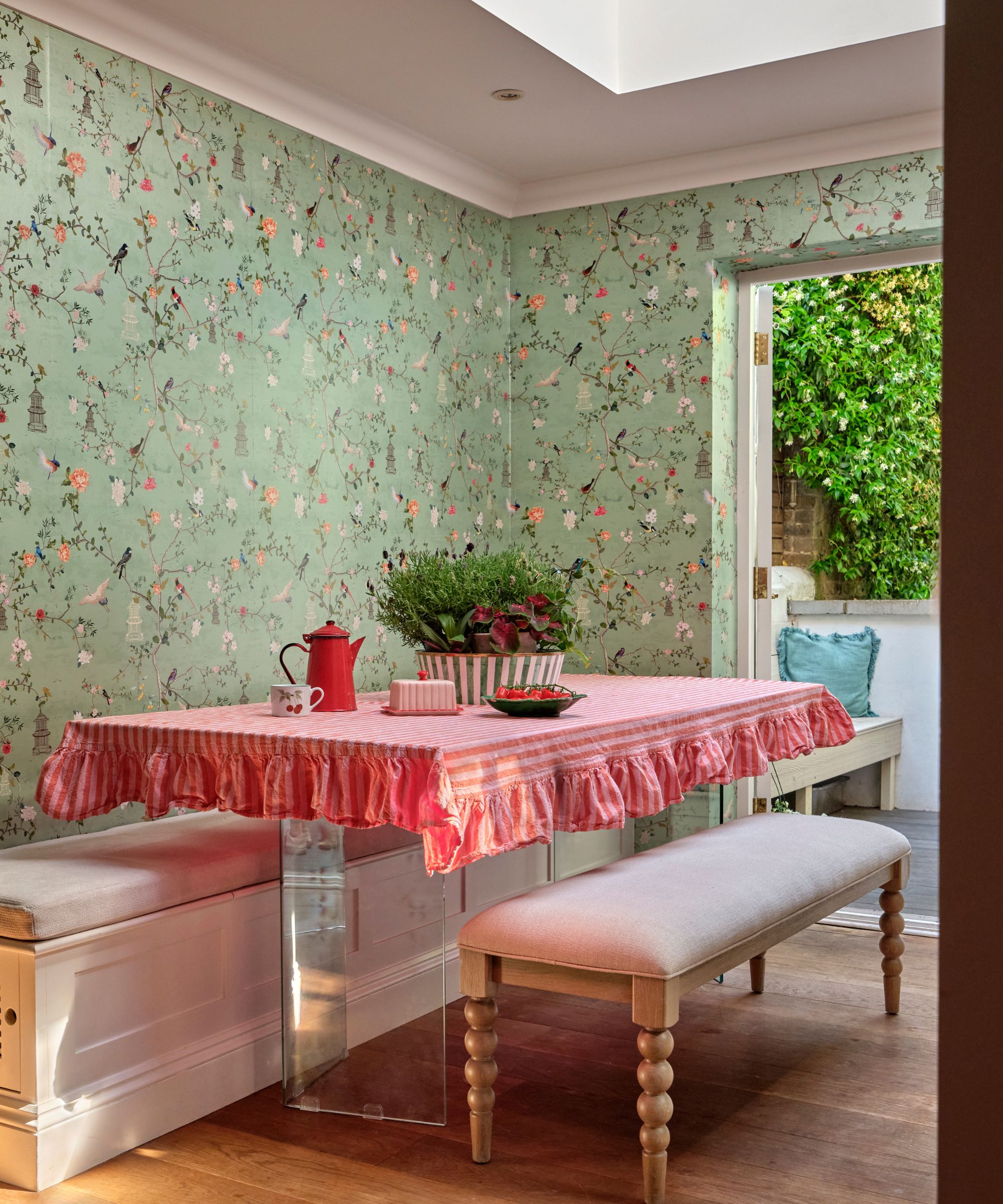 A dining area featuring a table covered in a ruffled pink striped cloth, flanked by built-in benches. The walls are covered in a pale green floral and bird motif wallpaper.