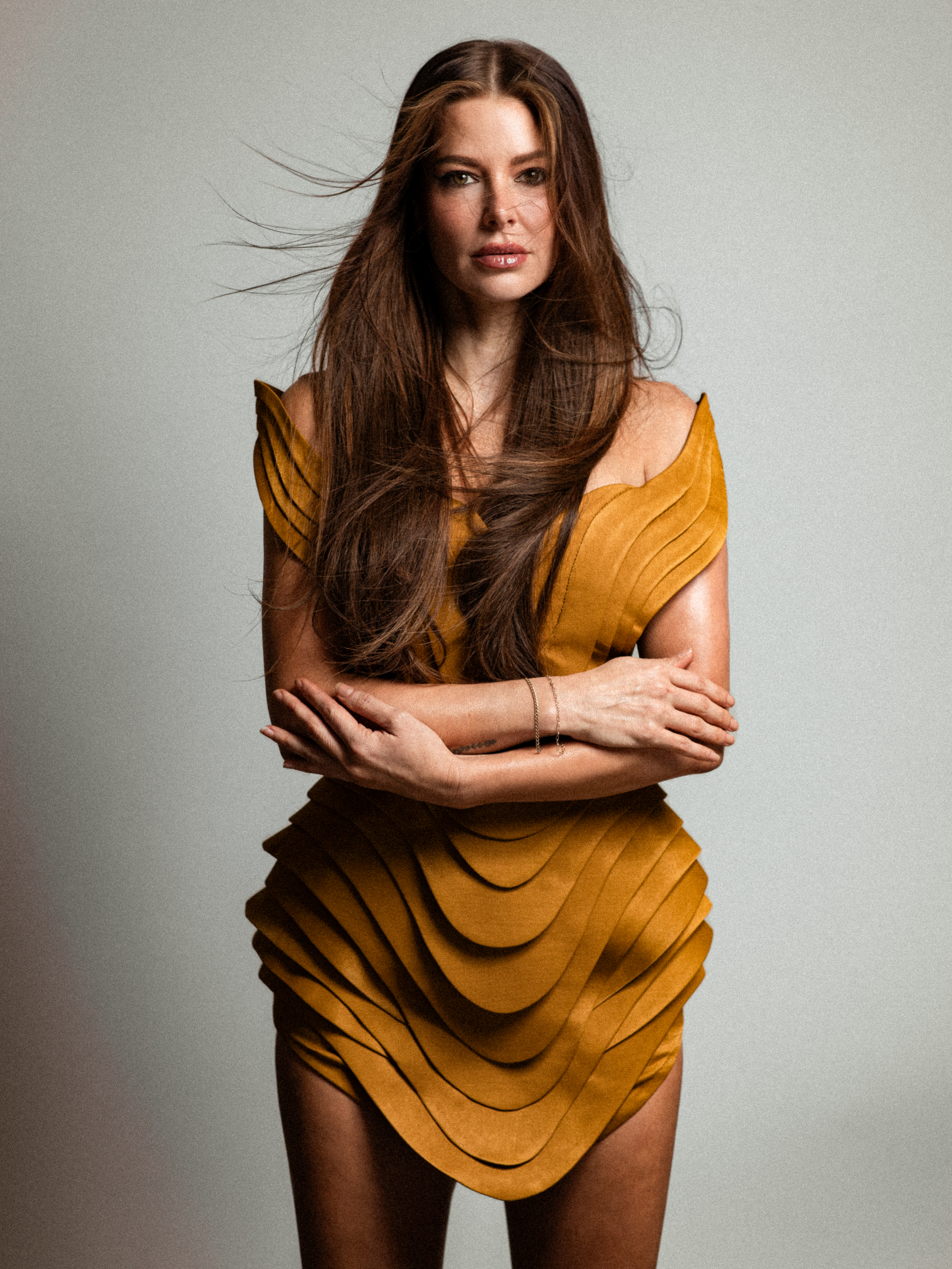 ariana madix wears a gold layered short dress and her brown hair down long and windswept as she poses crossing her arms