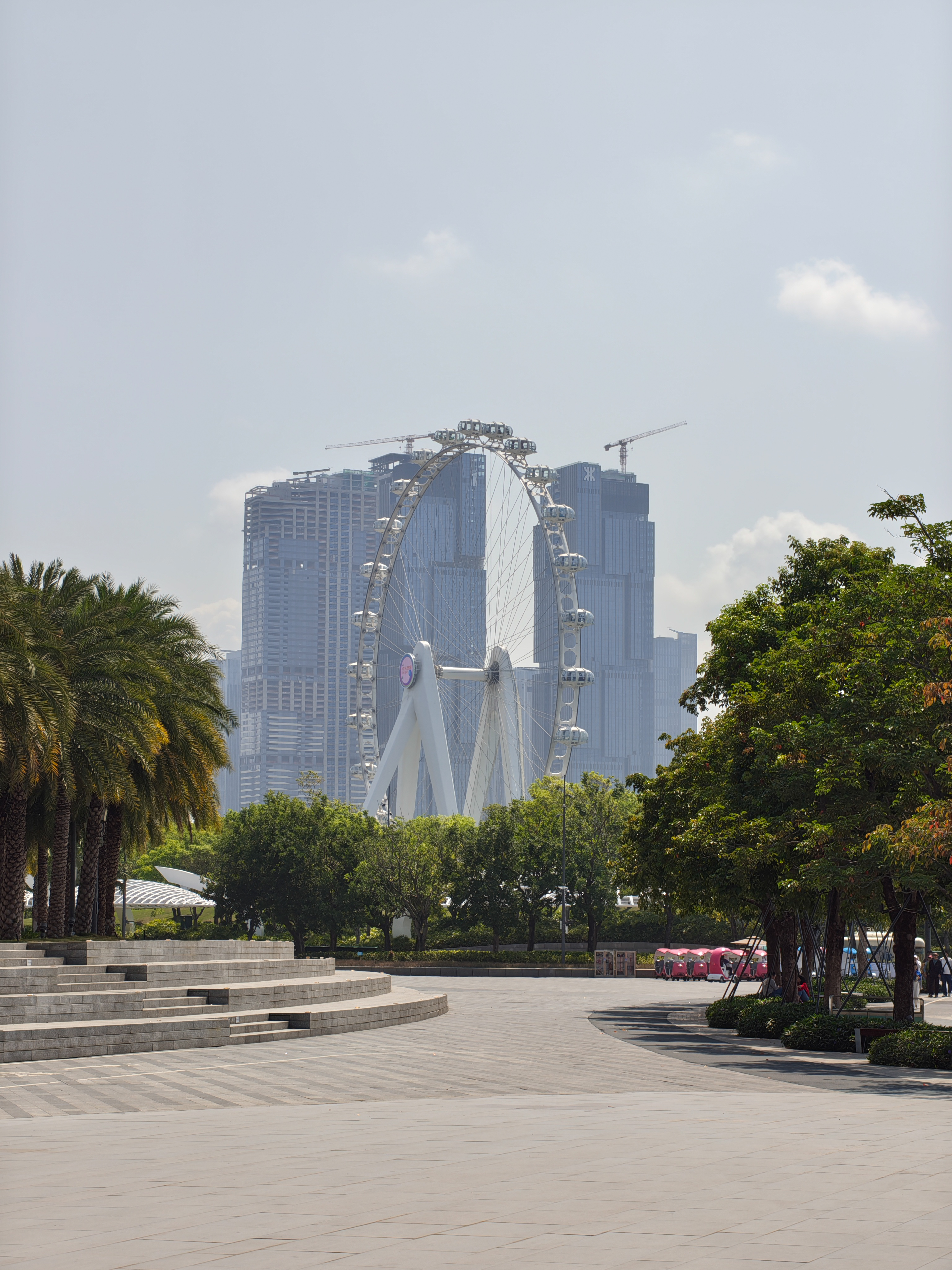 A medium shot of a large white Ferris wheel positioned in front of two tall, modern glass skyscrapers. The Ferris wheel is framed by green trees on both sides. In the foreground, a paved plaza features wide, low stone steps leading down from the left.