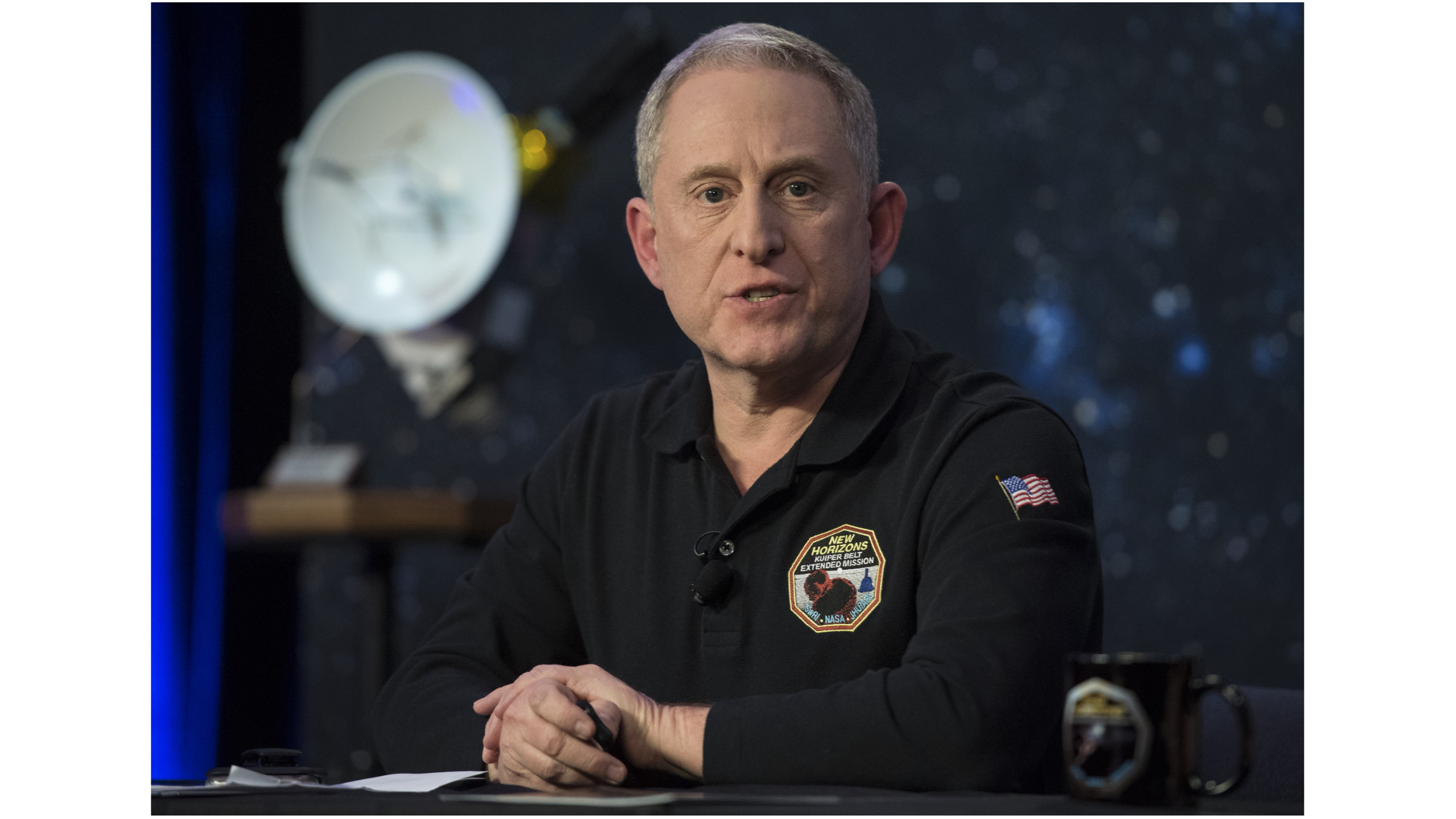 photo of a middle-aged man in a dark, long-sleeved shirt sitting at a table, with a model of a spacecraft in the background