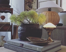 A stack of books, a houseplant, a wicker lamp with a white shade, and a brass candle holder holding a puck light