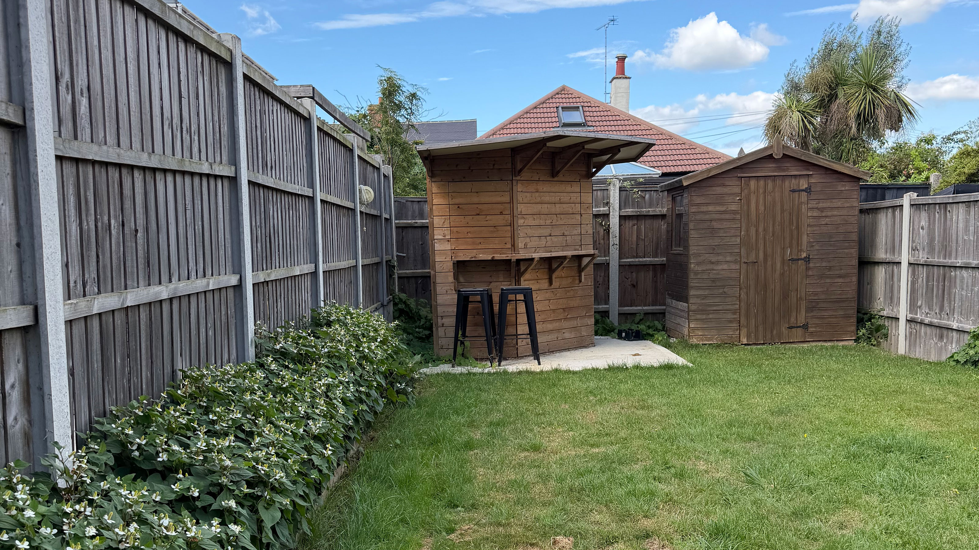 Lawn in garden surrounded by wooden fence and flower beds, with wooden garden bar and shed at end