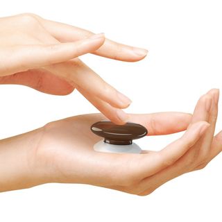 A brown Fibaro button is shown being held in one hand while a finger from another hand presses it.