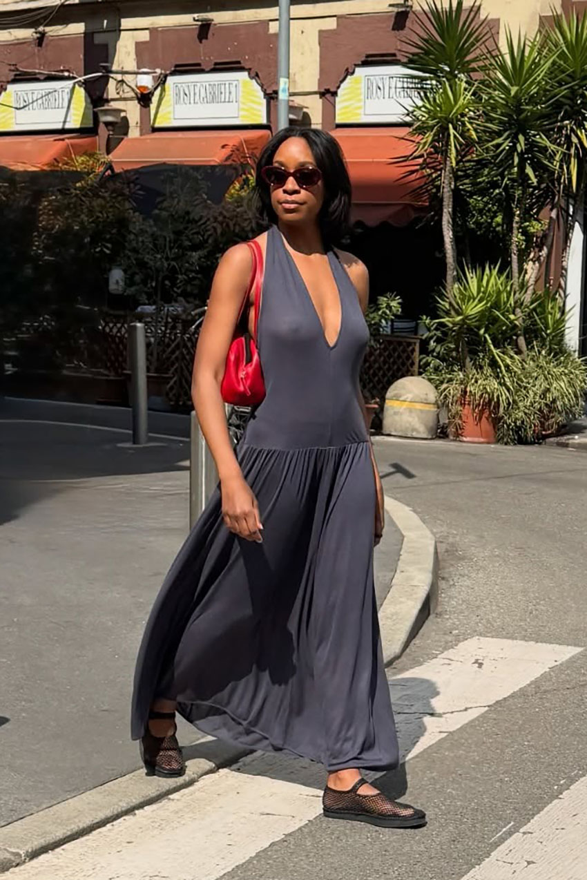 A woman wearing a gray deep plunge dress, red shoulder bag, and mesh flats.