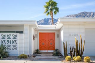 A modernist Californian house features paneled white walls and an orange geometrical front door, both of which stand out against the blue of the sky and a green, towering palm.